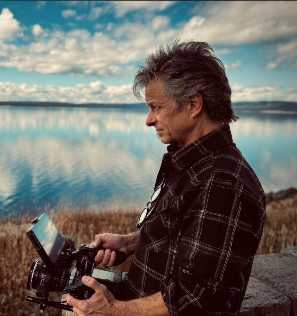 A man in a plaid shirt stands by a calm lake, holding a professional camera and monitor, with clouds and blue sky reflected in the water behind him.